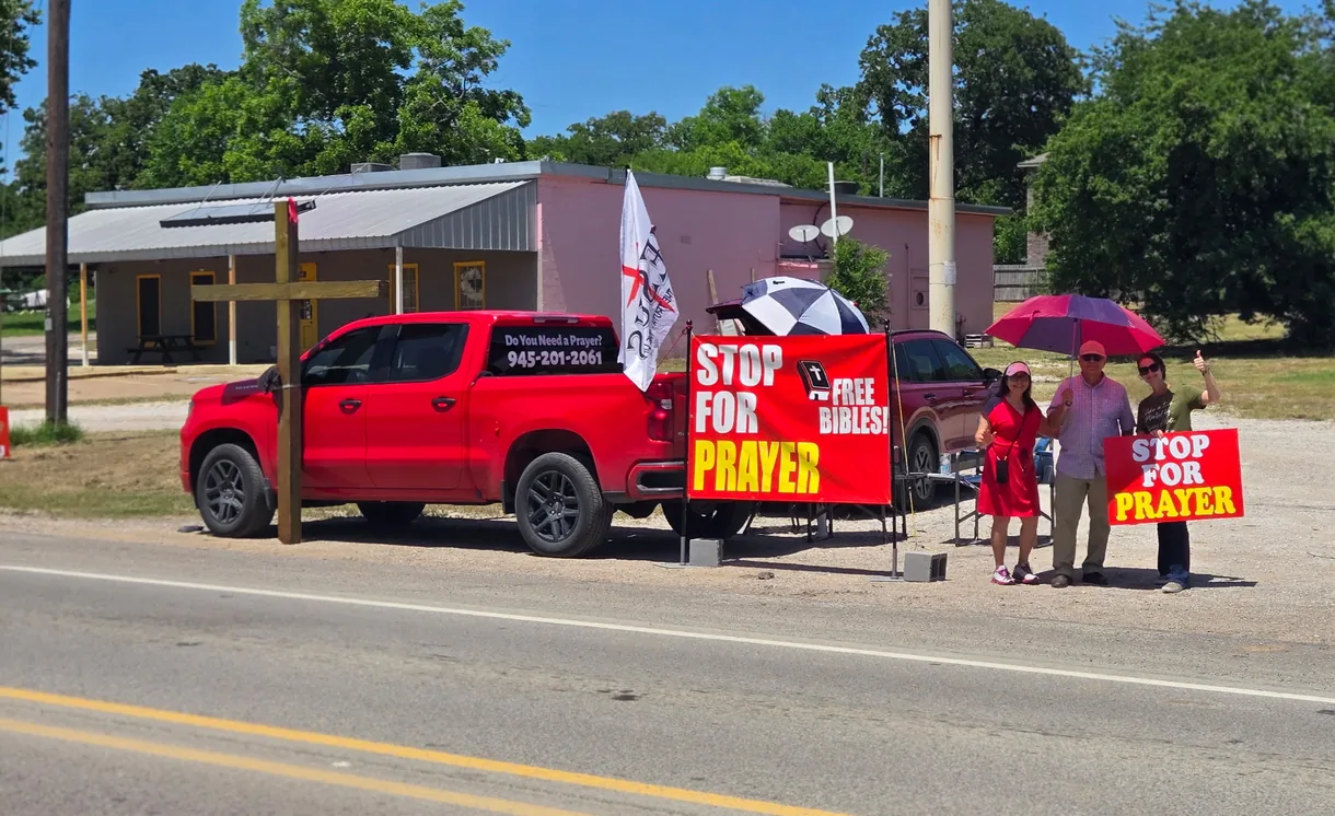 Wayne’s roadside prayer station — a red truck beside a wooden cross, Stop for Prayer banners offering free Bibles, and the team waiting under umbrellas.