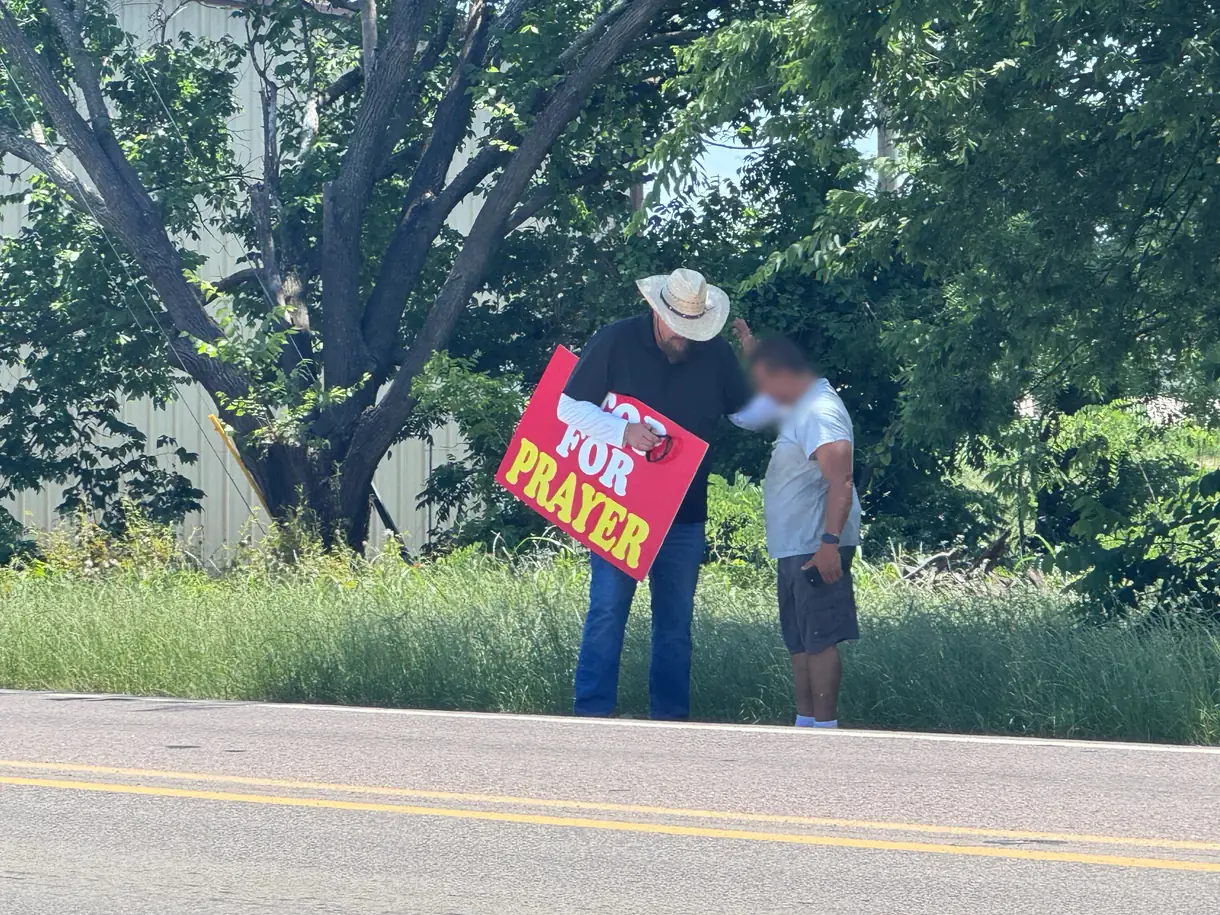 Wayne holding a Stop for Prayer sign at the roadside, praying with a man who has bowed his head.